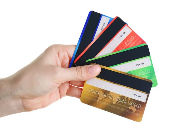 Mão segurando cinco cartões de crédito de cores diferentes — azul, vermelho, verde, dourado e preto — exibindo a faixa magnética no verso, sobre fundo branco.