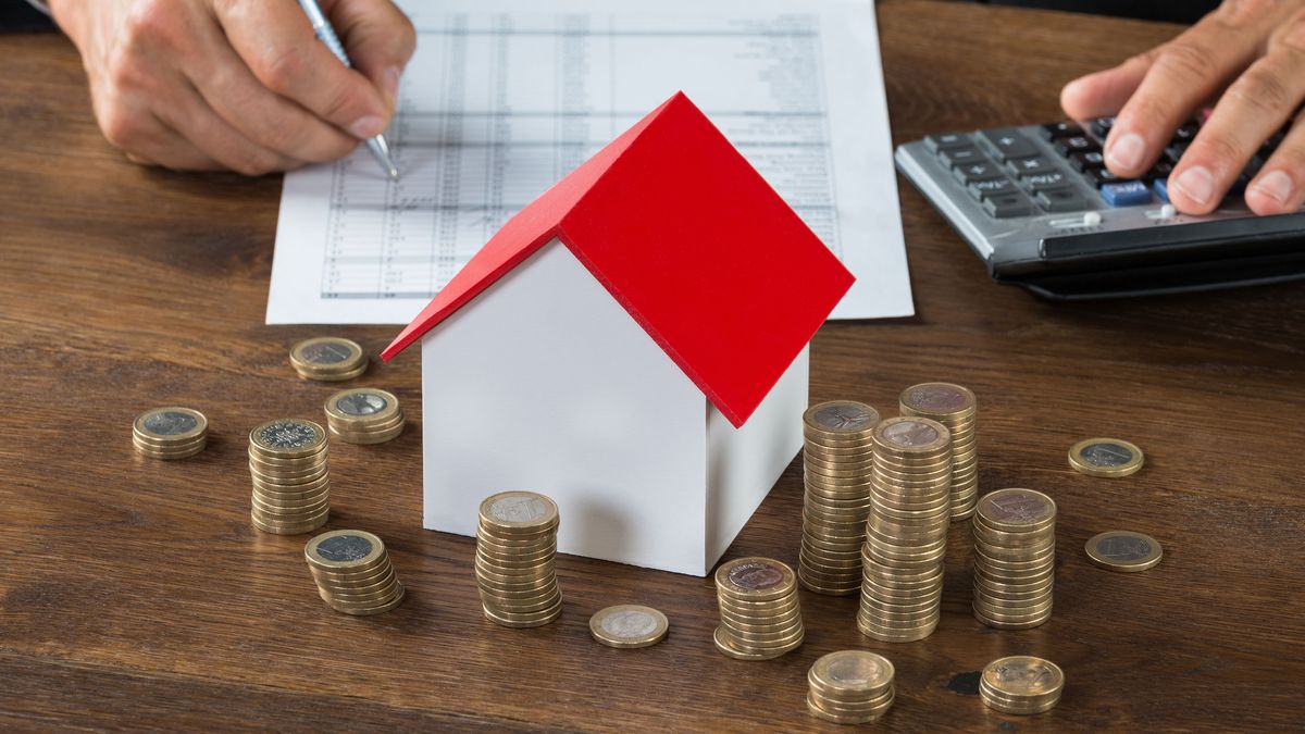 Casa en miniatura con techo rojo rodeada de pilas de monedas sobre una mesa de madera, mientras una persona realiza cálculos financieros, simbolizando la gestión y planificación de un préstamo hipotecario.